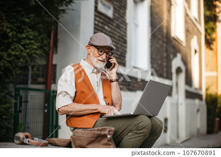 Senior man sitting on street curb working on laptop outdoors. Elderly man using digital technologies, working on a notebook. Concept of seniors and digital skills. 107670891