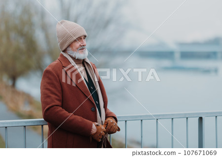Elegant senior man walking near a river, during cold day. Winter shot with copy space. 107671069