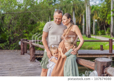 A happy, mature couple over 40 with their two daughters, enjoying a leisurely walk in a park, their joy evident as they embrace the journey of pregnancy later in life A happy, mature couple over 40 with their two daughters, enjoying a leisurely walk in a park, their joy evident as they embrace the journey of pregnancy later in life 107674080