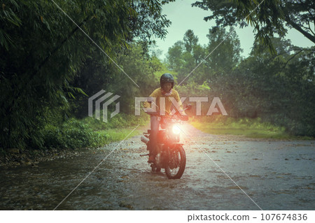man riding small endurom motorcycle crossing shallow creek among rain falling at forest 107674836