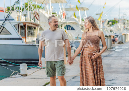 A happy, mature couple over 40, enjoying a leisurely walk on the waterfront, their joy evident as they embrace the journey of pregnancy later in life A happy, mature couple over 40, enjoying a leisurely walk on the waterfront, their joy evident as they embrace the journey of pregnancy later in life 107674928