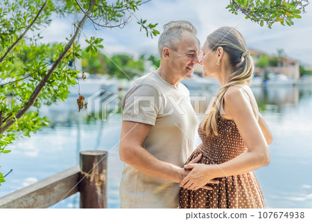 A happy, mature couple over 40, enjoying a leisurely walk on the waterfront, their joy evident as they embrace the journey of pregnancy later in life A happy, mature couple over 40, enjoying a leisurely walk on the waterfront, their joy evident as they embrace the journey of pregnancy later in life 107674938