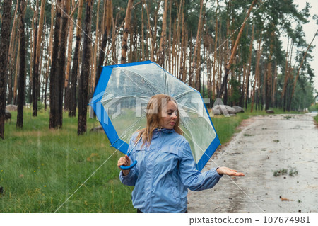 Blonde girl checking the weather under transparent umbrella outside in park. Autumn time rainy bad forecast conditions season. Tourist with umbrella travel in new places in rain Blonde girl checking the weather under transparent umbrella outside in park. Autumn time rainy bad forecast conditions season. Tourist with umbrella travel in new places in rain 107674981
