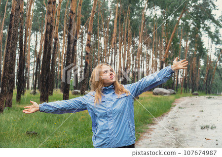 Happy young woman in blue raincoat enjoying the woods in park. True emotions open arms outdoors in rainy weather forecast. Tourist rest and feel freedom  107674987
