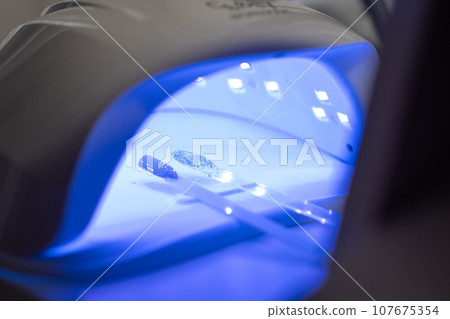 A manicurist applies gel nail polish to empty tips as a sample for future clients. Large range of gel polishes. Spa body care concept. LED nail lamp. Nail technician A manicurist applies gel nail polish to empty tips as a sample for future clients. Large range of gel polishes. Spa body care concept. LED nail lamp. Nail technician 107675354