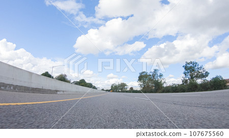 On a clear winter day, a car smoothly travels along Highway 101 near Santa Maria, California, under a brilliant blue sky, surrounded by a blend of greenery and golden hues. 107675560