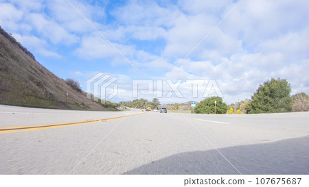 On a crisp winter day, a car cruises along the iconic Highway 101 near San Luis Obispo, California. The surrounding landscape is brownish and subdued, with rolling hills and patches of coastal 107675687