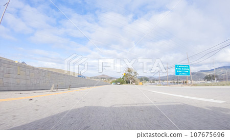 On a crisp winter day, a car cruises along the iconic Highway 101 near San Luis Obispo, California. The surrounding landscape is brownish and subdued, with rolling hills and patches of coastal 107675696