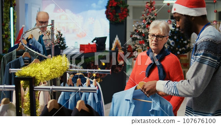 Worker assisting senior customer with picking right outfit to give as present to husband for upcoming xmas holiday season. Client browsing through clothes in festive ornate fashion shop 107676100