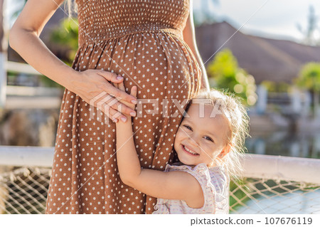 A heartwarming moment captured as a daughter lovingly embraces her mother's pregnant belly, sharing in the excitement and anticipation of a new family member 107676119