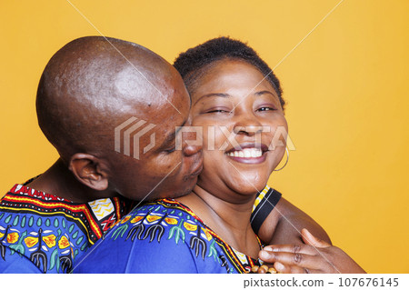Husband kissing smiling happy wife in cheek and hugging from back studio portrait. Cheerful african american woman looking at camera with carefree expression while embracing man 107676145