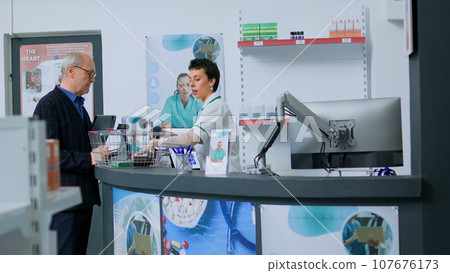 Pharmacy technician at drugstore counter scanning pharmaceutical products from elderly customer shopping basket. Older man buying sickness preventive medicaments in apothecary Pharmacy technician at drugstore counter scanning pharmaceutical products from elderly customer shopping basket. Older man buying sickness preventive medicaments in apothecary 107676173