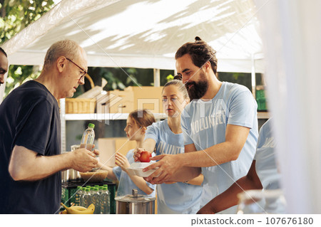 Friendly voluntary individuals helping the less fortunate, providing charitable support through food distribution. Young people volunteering and fighting poverty at an outdoor hunger relief program. 107676180