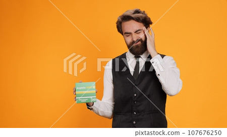Classy waiter with headache recommends pills on camera, showcasing pack of painkillers to ease pain. Young professional worker feeling unwell and ill, holding medicine treatment box. Classy waiter with headache recommends pills on camera, showcasing pack of painkillers to ease pain. Young professional worker feeling unwell and ill, holding medicine treatment box. 107676250