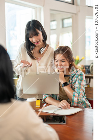Two pretty Asian female friends are working on a laptop while having a meeting with their team. Two pretty Asian female friends are working on a laptop while having a meeting with their team. 107676501