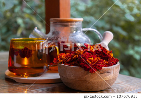 Marigold tea still life on table in green garden background. Healthy hot drink benefits. Natural organic aromatic drink in cup. Home-grown immunity-boosting herbs for tea. Autumn winter warming drink Marigold tea still life on table in green garden background. Healthy hot drink benefits. Natural organic aromatic drink in cup. Home-grown immunity-boosting herbs for tea. Autumn winter warming drink 107676913