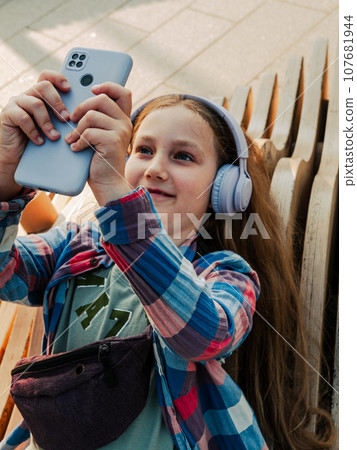 Blonde child in headphones listening to music podcast smartphone outside Young girl learn online lesson enjoy audio book 107681944
