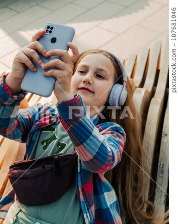 Blonde child in headphones listening to music podcast smartphone outside Young girl learn online lesson enjoy audio book 107681946