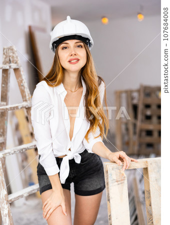 Sensual long-haired woman in hardhat, shirt and shorts posing in room under renovation 107684008