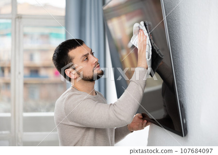 Young man wiping dust on screen of TV in living room Young man wiping dust on screen of TV in living room 107684097