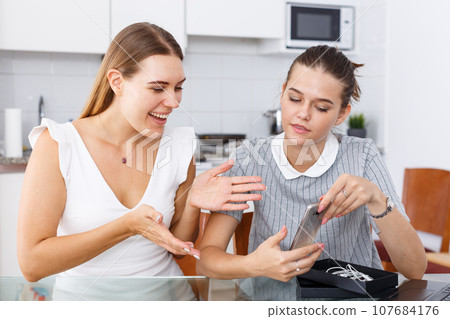 Young smiling woman showing new smartphone from box to female friend Young smiling woman showing new smartphone from box to female friend 107684176