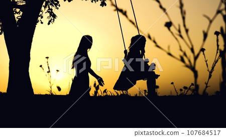 Mother riding little girl on swings dark silhouettes in park at sunset light. Little girl playing swings with mother silhouettes at dusk. Family of mother and daughter play swings in summer evening 107684517