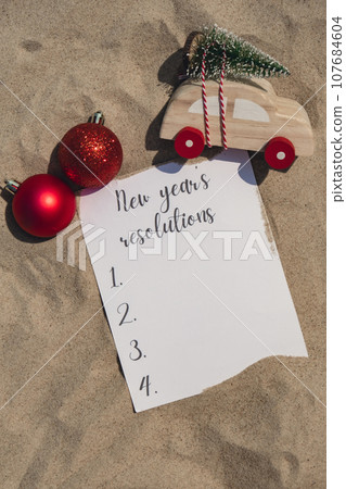 NEW YEAR RESOLUTIONS text on paper greeting card on background sandy beach sun coast. Christmas balls Santa hat New Year New Me goals decoration. Summer vacation decor. Holiday concept calendar date 107684604