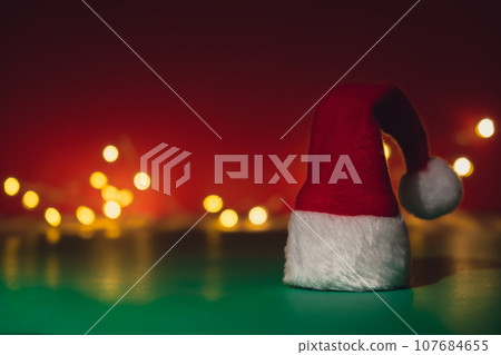 Santa hat on isometric green red background. Defocused lights on backdrop Copy space for your text. Mock up for advertisement. Holiday Christmas composition. Minimalistic greeting card 107684655