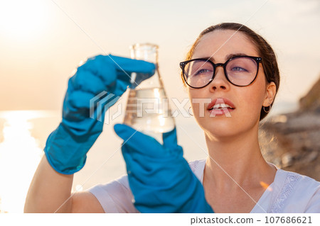 Portrait of ecologist wearing protective gloves and eyeglasses, examines test sample flask of water. Concept of pollution water and ecology Portrait of ecologist wearing protective gloves and eyeglasses, examines test sample flask of water. Concept of pollution water and ecology 107686621