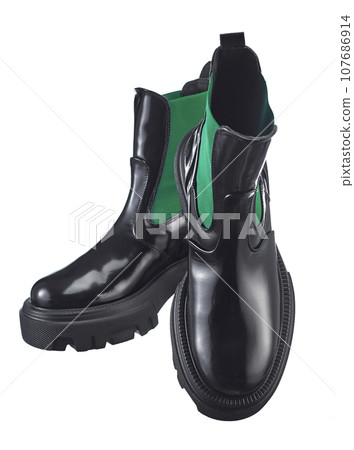 Beautiful pair of high demi-season boots made of glossy black leather with a green elastic band on the sides, with a massive sole, isolated on a white background. Beautiful pair of high demi-season boots made of glossy black leather with a green elastic band on the sides, with a massive sole, isolated on a white background. 107686914