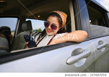 Pretty young woman leaning on car window and using mobile phone during travel nature road trip Pretty young woman leaning on car window and using mobile phone during travel nature road trip 107687059