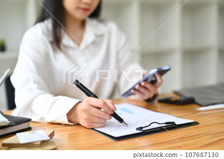 Female secretary holding mobile phone and writing appointments, schedule and business contacts at desk Female secretary holding mobile phone and writing appointments, schedule and business contacts at desk 107687130