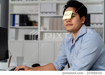 Young adult Asian businessman having HELP sticky note on his face in the office setting background. Business man is bored, frustrated, unhappy, and workaholic. 107688191
