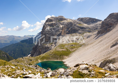 White sandy-rocky slope descends to shore of small lake in Laghi dei piani area with emerald water 107689808