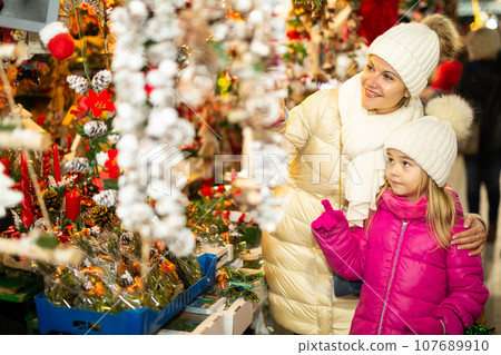 Smiling female and her daughter are choosing decorations for Christmas tree in the market outdoor Smiling female and her daughter are choosing decorations for Christmas tree in the market outdoor 107689910