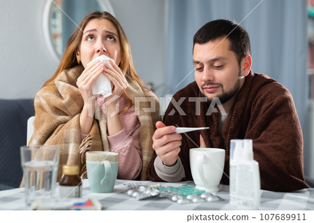 Couple suffering from flu and cough sitting wrapped in blankets Couple suffering from flu and cough sitting wrapped in blankets 107689911