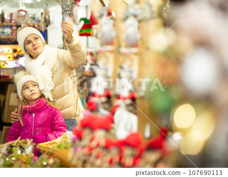 Smiling female and her daughter are choosing decorations for Christmas tree in the market outdoor Smiling female and her daughter are choosing decorations for Christmas tree in the market outdoor 107690113