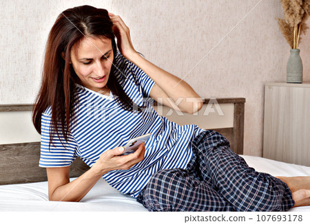 Brunette woman sitting in white bed using smartphone, having video call, smiling young woman relaxing in bedroom, waking up in the morning and sending text messages on one device, technology concept. Brunette woman sitting in white bed using smartphone, having video call, smiling young woman relaxing in bedroom, waking up in the morning and sending text messages on one device, technology concept. 107693178