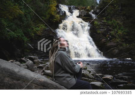 young beautiful Asian woman with African braids against the backdrop of a picturesque waterfall surrounded by forest 107693385