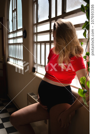 young pregnant woman hugging her big cute tummy while standing by the big window in vintage apartment. the concept of self-love and harmony young pregnant woman hugging her big cute tummy while standing by the big window in vintage apartment. the concept of self-love and harmony 107693386