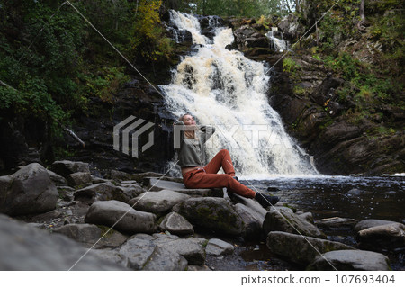 a young beautiful woman tourist sitting against the backdrop of a waterfall surrounded by forest a young beautiful woman tourist sitting against the backdrop of a waterfall surrounded by forest 107693404