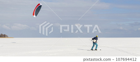 Panoramic view of many people friends enjoy riding kite surf board in warm suit on bright sunny winter day at frozen lake field snowy surface. Wintersport adrenaline fun adventure hobby acitivity 107694412
