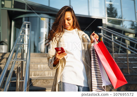 Young woman standing along road holding shopping bags, using mobile phone. Discounts, sale concept. 107695572