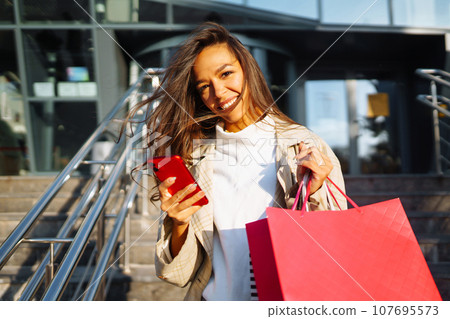 Young woman standing along road holding shopping bags, using mobile phone. Discounts, sale concept. 107695573