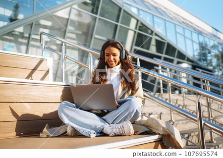 Young woman in headphones with laptop on city street. Video conference. ?nline training. Freelancer 107695574