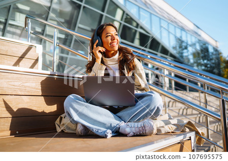 Young woman in headphones with laptop on city street. Video conference. ?nline training. Freelancer 107695575