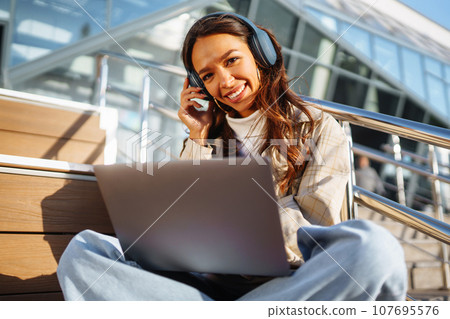 Young woman in headphones with laptop on city street. Video conference. ?nline training. Freelancer 107695576