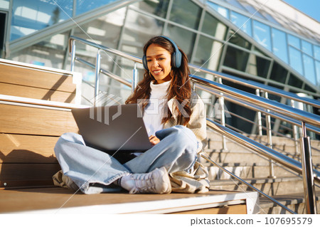 Young woman in headphones with laptop on city street. Video conference. ?nline training. Freelancer 107695579