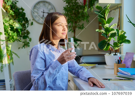 Young woman drinks water from glass, sitting at desk with laptop at home Young woman drinks water from glass, sitting at desk with laptop at home 107697039