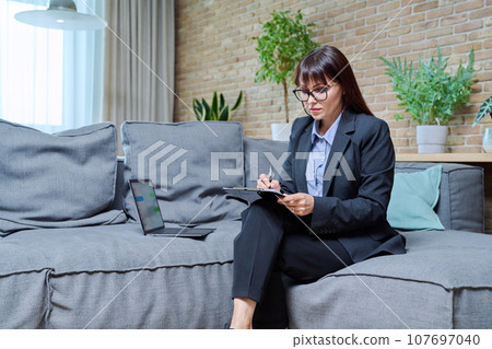 Portrait of female psychologist with clipboard sitting on couch in office Portrait of female psychologist with clipboard sitting on couch in office 107697040
